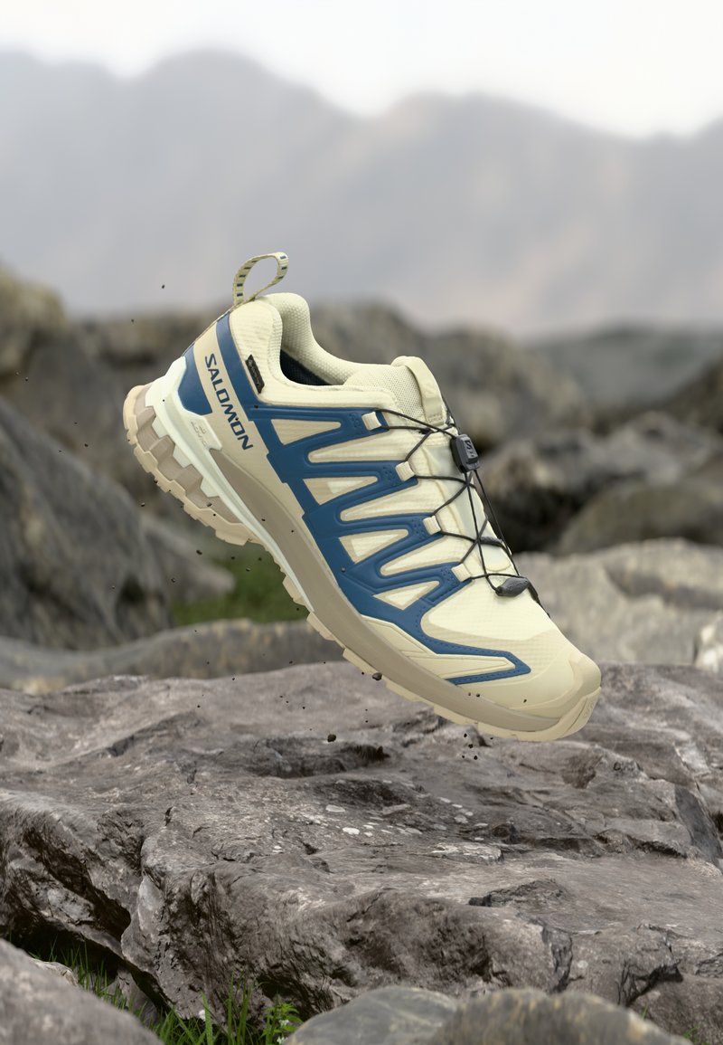 Leichter beiger Trailrunning-Schuh mit blauen Akzenten, strukturiertem Mesh-Obermaterial und einer robusten Sohle, ausgestattet mit elastischen Schnürsenkeln und einer Zuglasche.