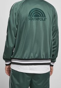 Grüne Satin-Bomberjacke mit einem schwarz-weißen Streifenbund, einem großen grünen und schwarzen "SOUTHPOLE"-Logo und einem weißen Unterhemd.
