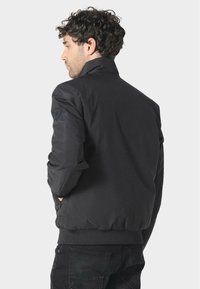 Czarna bomberka z wysokim kołnierzem, teksturowanym materiałem, elastycznymi mankietami i dołem; posiada naszywkę z logo na lewym rękawie.