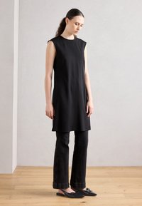 Vestido negro sin mangas con corte recto, llevado sobre pantalones de mezclilla oscuros acampanados; presenta costuras mínimas y zapatos planos negros puntiagudos.