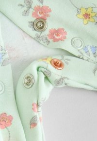 Tissu en coton vert clair avec des motifs floraux en rose et jaune, comportant des boutons-pression en argent, blanc et cuivre pour la fermeture.