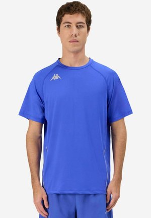 Homme portant un t-shirt de sport bleu uni à manches courtes et un short bleu assorti, debout devant un fond blanc.