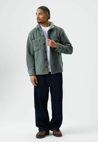 Groen, pluizig jasje met knopen en een borstzak, gedragen over een wit T-shirt, gecombineerd met donkerblauwe, losvallende broek en bruine schoenen.