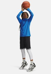Maglietta sportiva a maniche lunghe blu, pantaloni corti neri con dettagli bianchi, leggings bianchi e sneakers neri mentre tiene un pallone da basket.