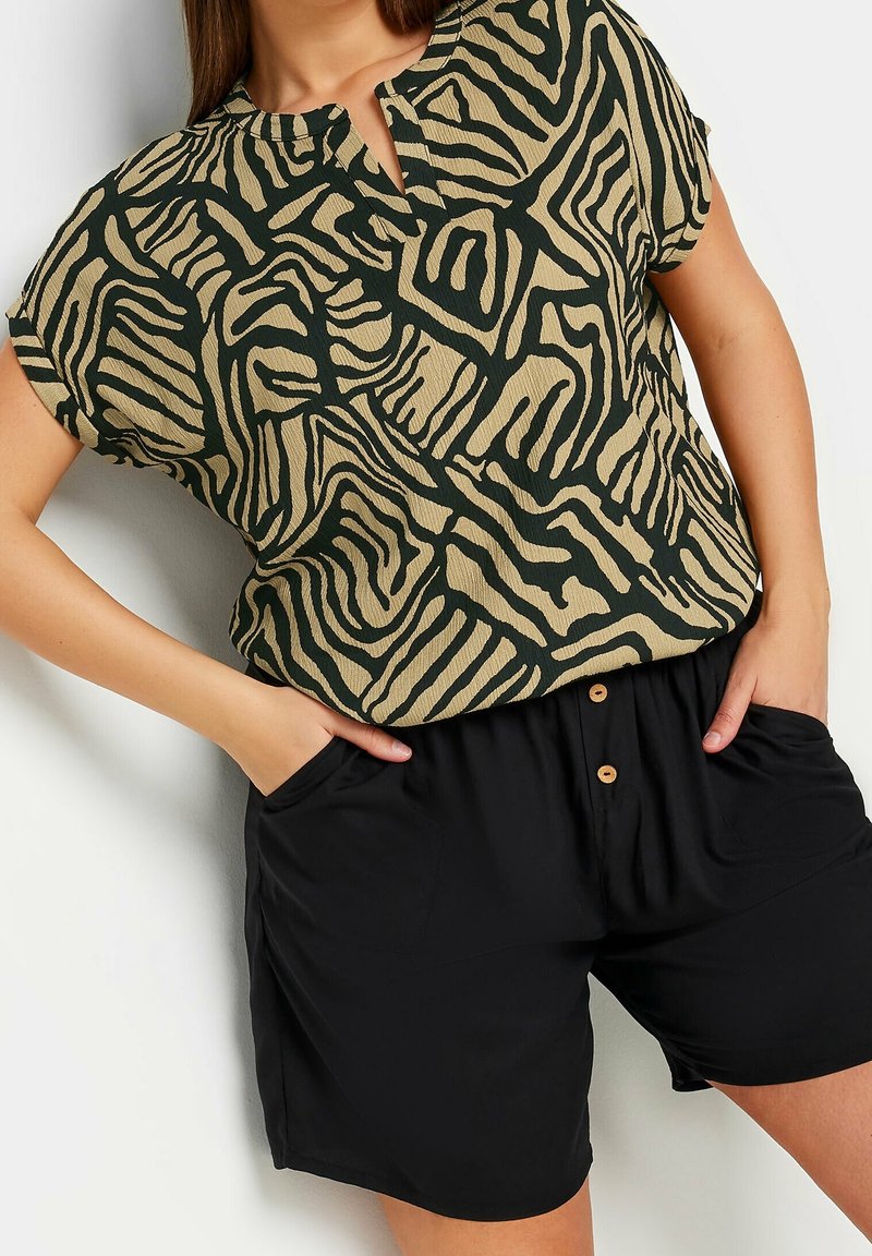 Blusa a maniche corte color sabbia con un motivo astratto a zigzag verde scuro, con scollatura a V. Abbinata a pantaloni corti neri con tasche.