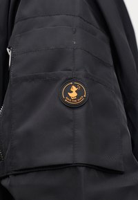 Poche de veste noire avec un patch rond orange représentant un canard marchant et le texte "SAVE THE DUCK" à l'intérieur de flèches circulaires.