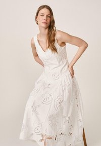 Mujer con cabello largo, vestida con un vestido blanco sin mangas con bordados de motivos marinos recortados, posando con las manos en las caderas contra un fondo liso.