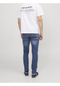 Man wearing white graphic T-shirt and blue jeans standing facing away against plain background.