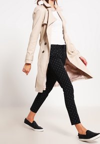 Beige trench coat s dvojitými knoflíky, nošený přes bílý top a vzorované černé skinny kalhoty. Černé nazouváky dokončují outfit.