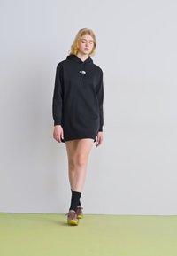 Vestido moletom preto com mangas longas, apresentando um capuz com cordão. A modelo usa meias pretas e sapatos castanhos com sola amarela em um chão verde.