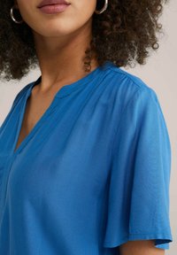 Blå V-ringad tröja gjord av lättviktsmaterial; har korta ärmar, mjuk textur och subtila veck längs halsringningen.