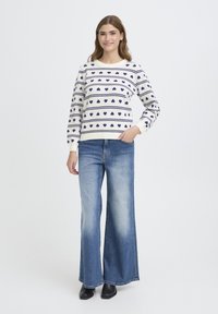 Pull crème avec motifs de cœur et rayures bleu marine, associé à un jean à jambes larges bleu clair. Le modèle pose avec une main dans la poche.