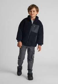 Veste en polaire noire avec un col haut, fermeture éclair et une poche contrastante. Un pantalon cargo gris et des baskets noires complètent la tenue.
