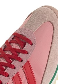 Rózsaszín sportcipő vörös fűzőkkel, velúr és textil felsőrésszel, zigzag díszítéssel, párnázott bokarésszel, valamint zöld és fehér masszív gumitalppal.