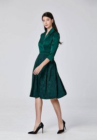 Femme portant une robe longue noire, arrivant aux genoux, à manches trois-quarts et des talons hauts noirs, se tenant devant un fond clair.