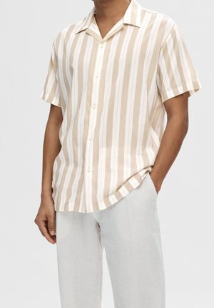 Homme portant une chemise boutonnée à manches courtes beige et blanche à rayures verticales avec un pantalon de couleur claire sur un fond uni.