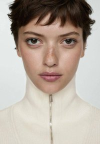 Närbild av en person som bär en krämfärgad höghalssweater med en synlig silverdragkedja, med ljusa fräknar och neutral smink.