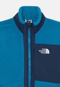 Flis jakna blagovne znamke The North Face z modro teksturo, visokim ovratnikom, sprednjim zadrgo, mornarsko modrimi detajli in logotipom na levem prsnem delu.