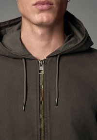 Gros plan d'une personne portant un sweat à capuche marron zippé avec des cordons, montrant le cou et le bas du visage, lèvres fermées, expression neutre.