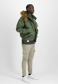 Grüne Bomberjacke mit Kunstfellkapuze, dazu ein dunkler Pullover, Cargo-Hose und weiße Sneakers. Verfügt über einen Reißverschluss und einen Druckknopfverschluss.