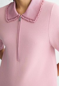 Haut zippé rose clair en tissu texturé, doté d'un col perlé composé de perles rondes et d'accents métalliques. Manches courtes.