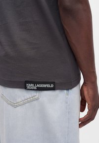 Μαύρο τελείωμα πουκάμισου με ετικέτα "Karl Lagerfeld Jeans", φορεμένο πάνω από ανοιχτόχρωμα τζιν σε άτομο με σκούρο δέρμα.
