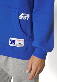 Poche de sweat-shirt bleu avec texte blanc "depuis 1937" et écusson Mitchell & Ness, porté avec un pantalon de survêtement gris et une main visible.
