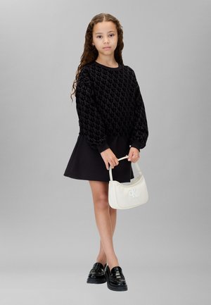 Pull noir à motifs avec des manches ballon, associé à une jupe noire évasée. Tenant un sac à main blanc avec un logo de marque. Chaussures noires à enfiler.