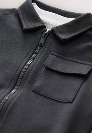 Zwarte zip-up jas met een kraag, gestructureerde stof, een borstzak en een contrasterende witte binnenkraag.