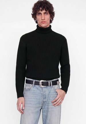 Un homme aux cheveux bouclés porte un pull noir à col roulé côtelé, un jean bleu clair et une ceinture noire à boucle argentée sur fond blanc.
