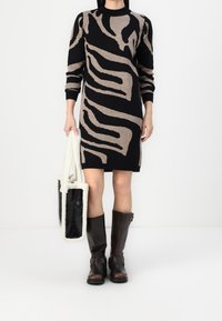 Femme portant une robe-pull à motif abstrait noir et beige, tenant un sac fourre-tout noir et blanc, et portant des bottes brun foncé montant jusqu'aux genoux.
