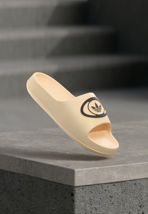 Jasnożółta klapka na płaskiej podeszwie wykonana z miękkiego materiału, z otwartym noskiem, czarnym emblematem logo na górnej części oraz gładką fakturą.