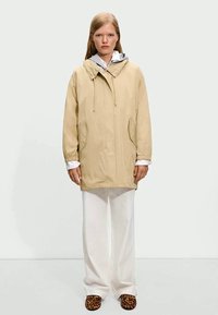 Μπεζ μπουφάν με κουκούλα και κορδόνια, πλευρικές τσέπες και ελαστικές μανσέτες, φορεμένο πάνω από ένα λευκό πουκάμισο με μακριά μανίκια και συνδυασμένο με πλατιά λευκά παντελόνια και παπούτσια με σχέδια.