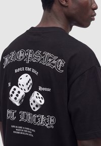 Homme portant un t-shirt noir avec un texte blanc et trois graphiques de dés dans le dos, incluant les phrases «DROP SIZE», «ROLL THE DICE» et «GET LUCKY».