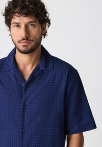 Korte mouw overhemd in donker marineblauw, gemêleerde stof met een subtiel geometrisch patroon. Voorzien van een gespreide kraag en knoopsluiting.