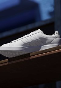 Hvid læder sneaker med tekstureret sål og parallel syede striber, placeret på en træflade mod en sløret mørkeblå baggrund.