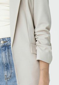 Blazer gris clair avec un tissu lisse et texturé, comprenant des détails froncés aux manches. Porté avec un jean bleu en denim, affichant un bouton et une poche.