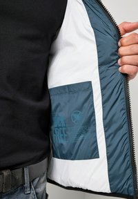 Giacca imbottita con interno turchese e bianco, chiusura con zip e una tasca con la scritta "Duck Free" in blu su materiale sintetico.