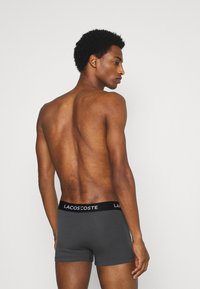 Boxers gris avec une ceinture noire arborant le logo "LACOSTE". Le matériau semble lisse avec un design ajusté, vu de l'arrière.