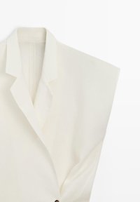 Blazer blanc sans manches avec col à revers cranté et fermeture à un bouton sur fond uni.