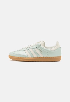 Světle modré tenisky Adidas Samba s bílými pruhy a gumovou podrážkou, zobrazené z bočního pohledu na jednoduchém pozadí.