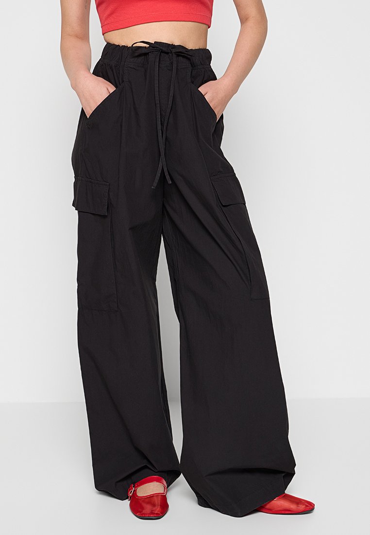 Pantaloni cargo neri a gamba larga con vita regolabile, due grandi tasche laterali e una consistenza morbida. Abbinati a scarpe basse rosse.