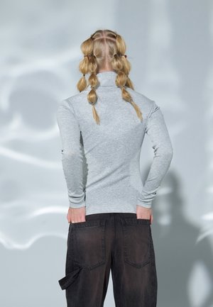 Personne aux cheveux blonds coiffés en plusieurs queues de cheval bulles, portant un haut à manches longues gris clair et un pantalon noir, les mains dans les poches arrière.