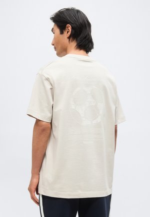 Homme aux cheveux foncés portant un t-shirt beige oversize avec un léger motif de ballon de football dans le dos, debout sur un fond uni.