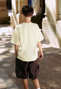 Light yellow short-sleeve t-shirt paired with dark brown cargo shorts, featuring side pockets, viewed from the back.