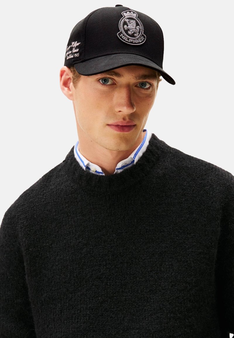 Gorra de béisbol negra con logotipo bordado y texto blanco, llevada por una persona con suéter negro y camisa con cuello con detalles en azul.