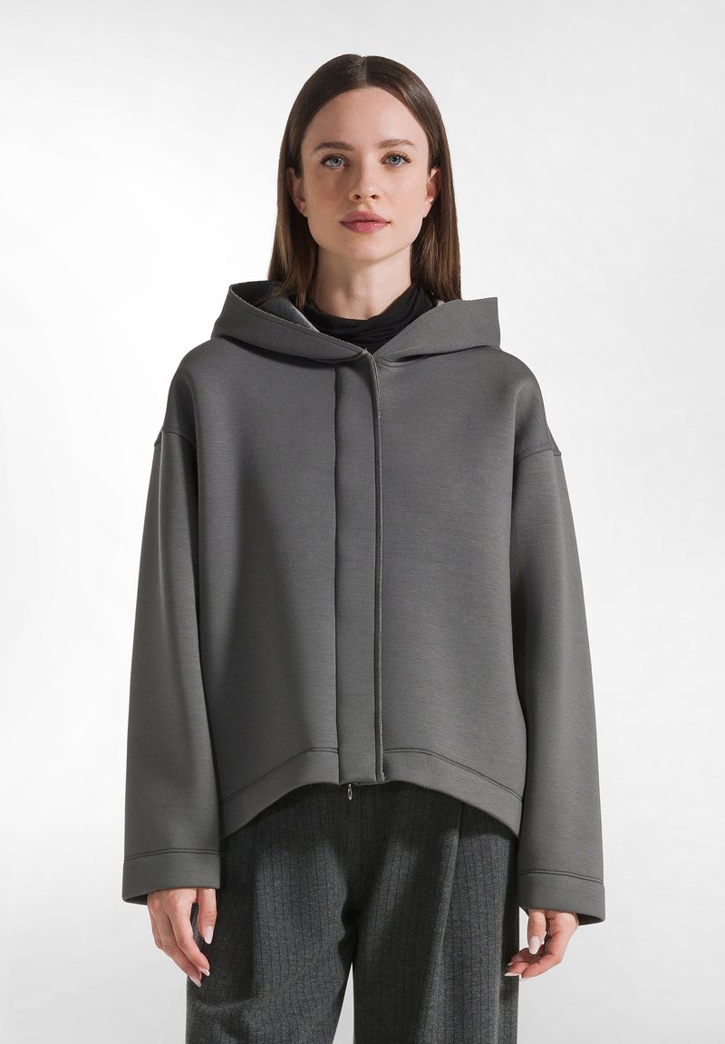 Felpa grigia con zip, dal taglio ampio, con cappuccio grande e rifiniture a costine. Texture liscia con una finitura leggermente lucida.