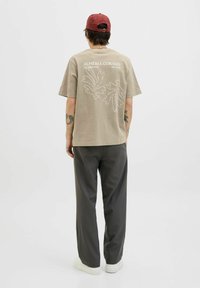 Beige T-Shirt mit weißem botanischen Grafikdruck auf der Rückseite, kombiniert mit grauen Hosen und weißen Sneakern. Kurze Ärmel, lockere Passform, minimales Design.