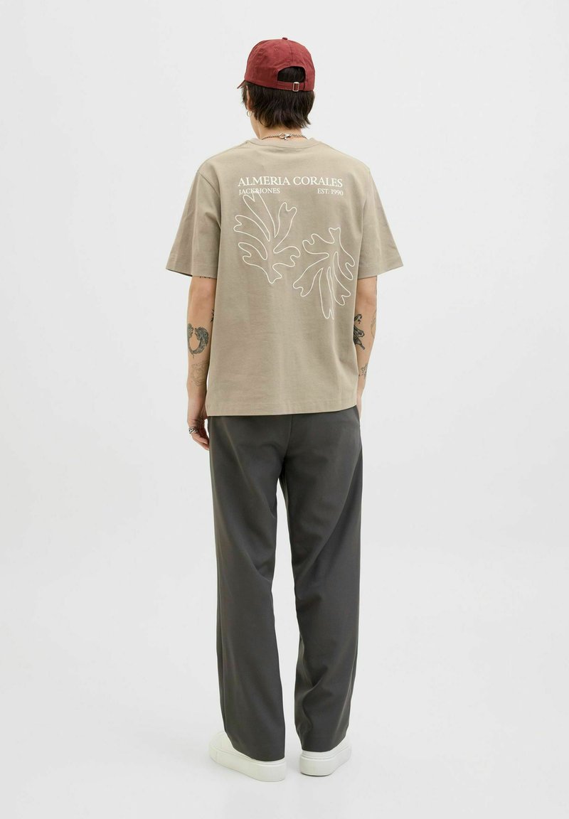 Beige T-Shirt mit weißem botanischen Grafikdruck auf der Rückseite, kombiniert mit grauen Hosen und weißen Sneakern. Kurze Ärmel, lockere Passform, minimales Design.