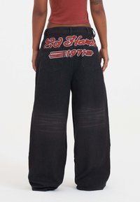 Osoba v širokých černých džínách se červeným a bílým vyšitím "Ed Hardy 1971" na zadních kapsách, stojící na bílém pozadí.